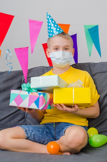 Il ragazzo felice nella maschera di protezione della medicina con i regali a disposizione celebra il compleanno