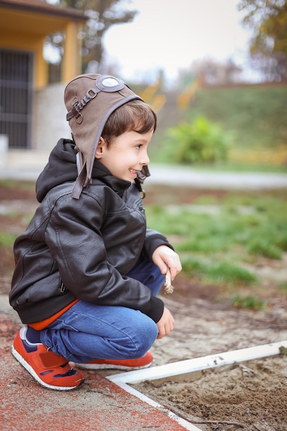 Happy boy in a pilot's cap and brown jacket plays with with sand in an autumn park and dreams.