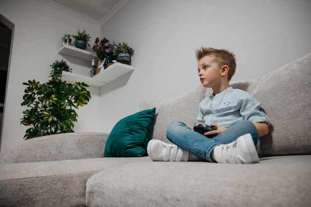 Happy boy playing video games holding game controller sitting\
on the coach in living room