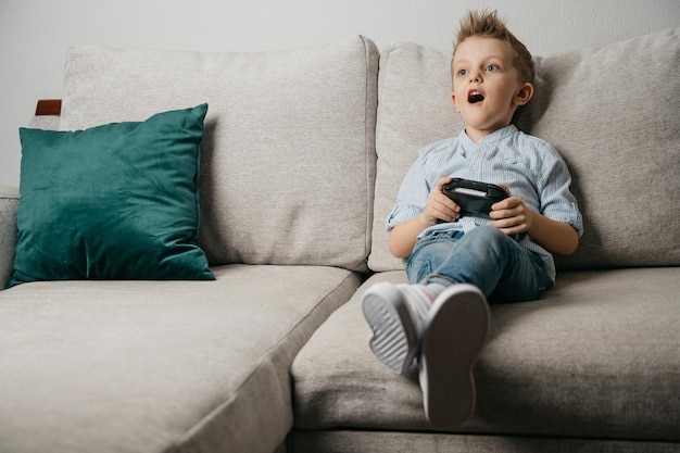 Happy boy playing video games holding game controller sitting\
on the coach in living room