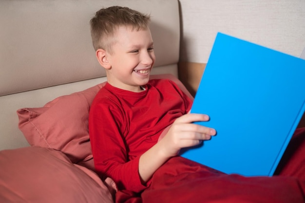 A happy boy in red pajamas is lying in bed and reading a book