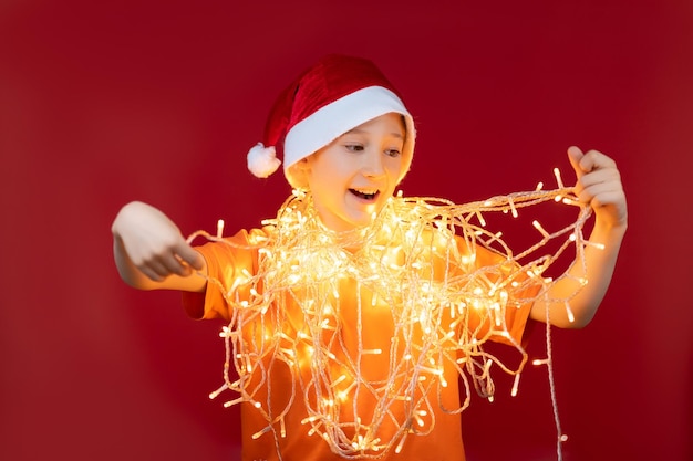 Happy boy in a red santa hat, entangled in a Christmas garland that glows
