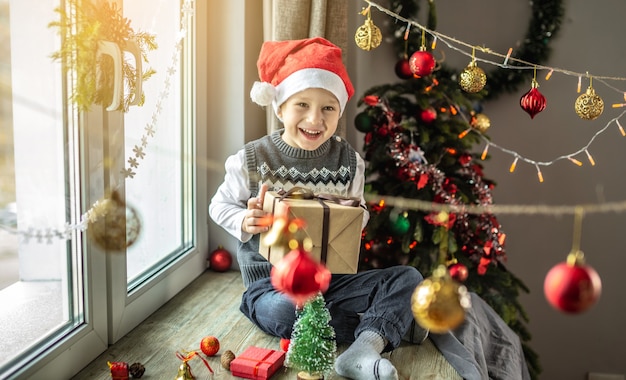 Happy boy in a red Santa hat is sitting at the window next to a Christmas tree in a room decorated for the holiday and holding a gift box in his hands. Concept of New Year atmosphere and festive mood.