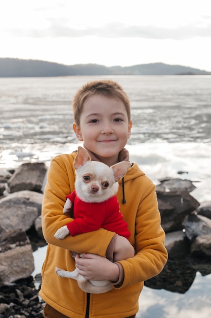 A happy boy on the river bank holds a Chihuahua dog in his arms and smiles. A child with a dog in nature.