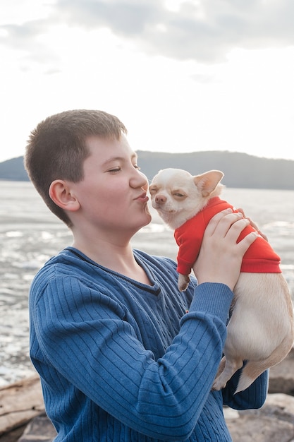 A happy boy on the river bank holds a Chihuahua dog in his arms and smiles. A child with a dog in nature.