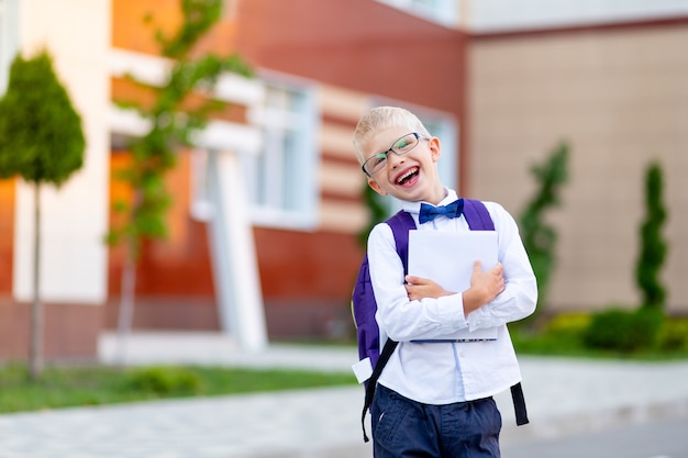 Happy boy schoolboy blond with glasses with a backpack and a white book stands at the school and laughs