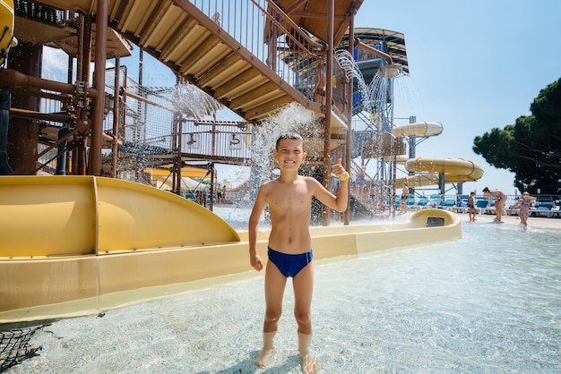 A happy boy of seven years old smiles and shows a class against the background of slides in a water park. Happy vacation vacation. Summer holidays and tourism.