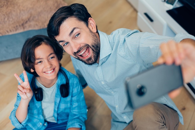 Happy boy showing peace gesture while an excited father taking photos with him at home