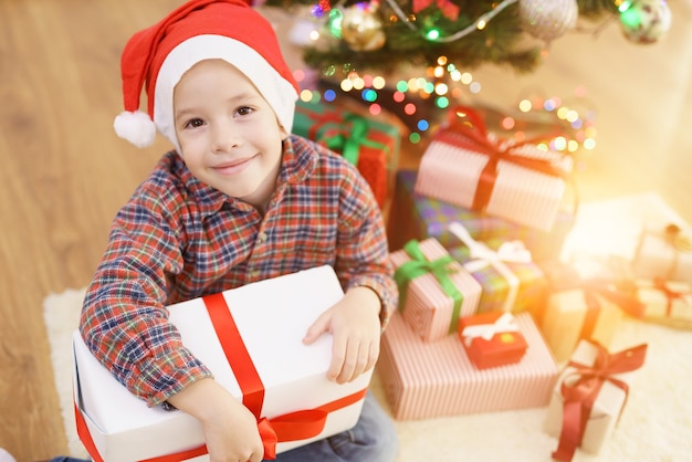 The happy boy sit with a gift box near the christmas tree on a sunny background