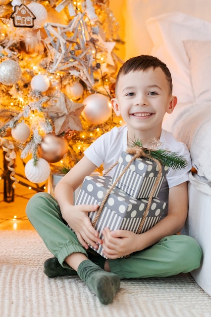 Happy boy sits on floor with gift in his hand by window in front christmas tree in his bedroom