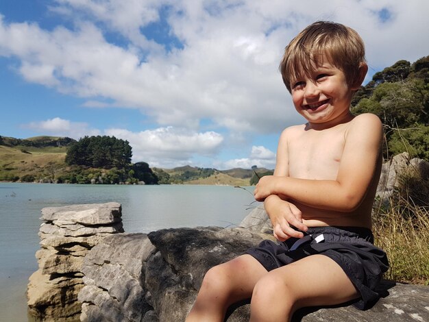 Photo happy boy sitting on rock by lake against sky
