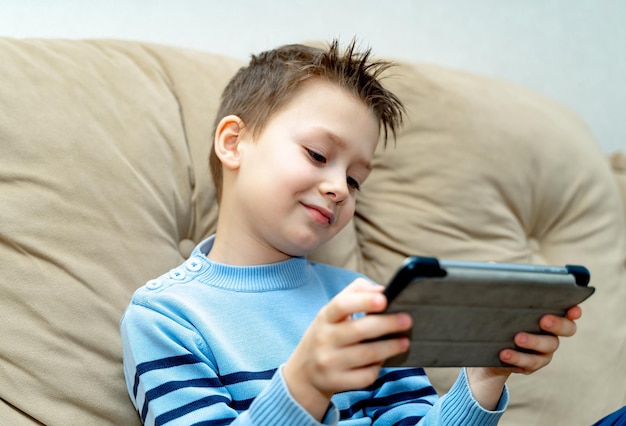 부드러운 소파에 앉아 집에서 최신 장치로 흥미로운 비디오를 보는 행복한 소년 디지털 태블릿을 사용하여 파란 스웨터에 표정이 풍부한 웃는 얼굴의 어린 소년