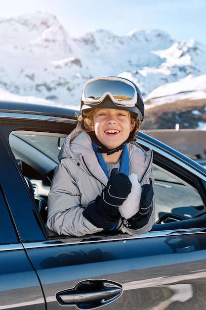 Happy boy in ski goggles leaning out window with snowball and looking at camera while riding on road against snowy mountains