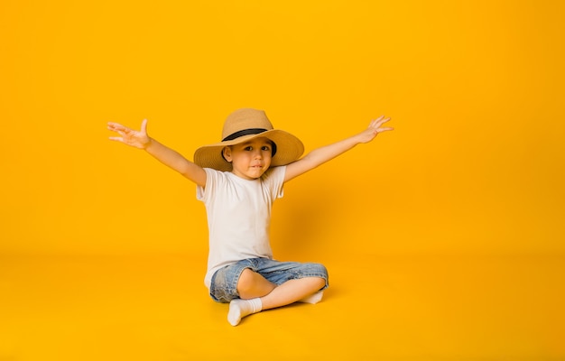 Happy boy in a straw hat sits on a yellow surface and points with his hands to the side