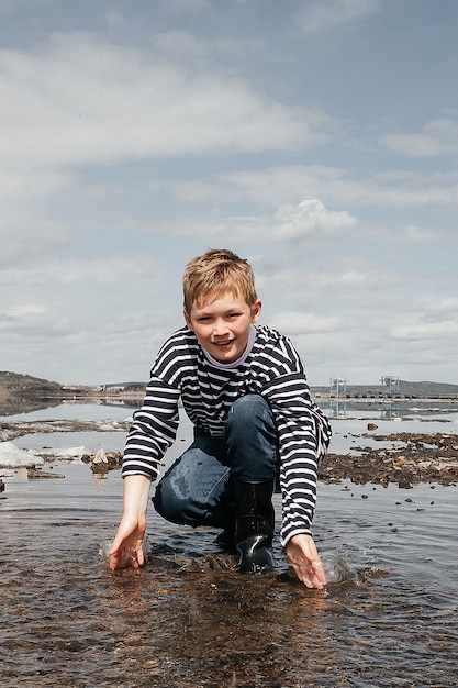 A happy boy in a striped vest and rubber boots on the bank of the river splashes water merrily. Outdoor recreation.