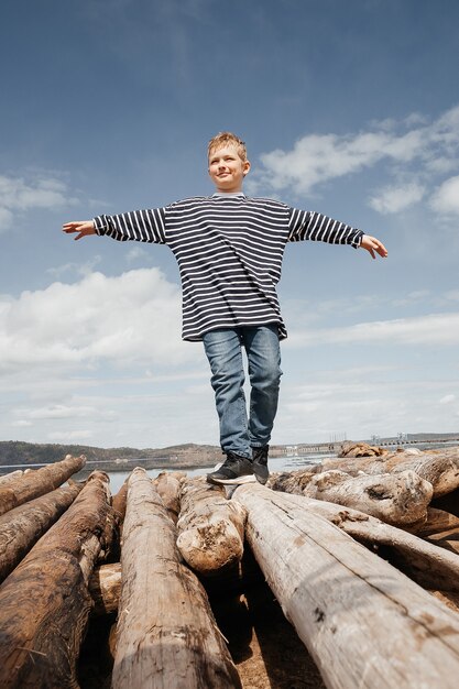 A happy boy in a striped vest walks along the logs to the shore. The boy tries to keep his balance as he walks over the logs. Weekend rest.