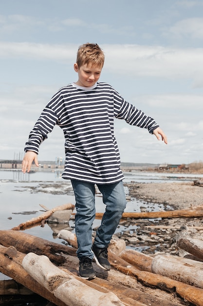 Photo a happy boy in a striped vest walks along the logs to the shore. the boy tries to keep his balance as he walks over the logs. weekend rest.