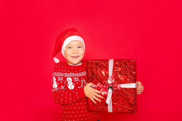 A happy boy in a sweater and Santa hat stands on an isolated red background with a large gift, space for text