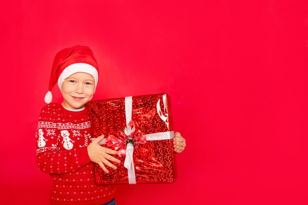 A happy boy in a sweater and Santa hat stands on an isolated red background with a large gift