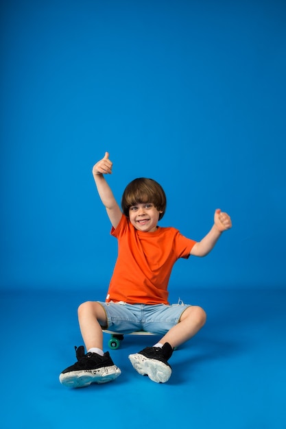 Happy boy toddler sits on a skateboard and holds his hands up on a blue wall with space for text