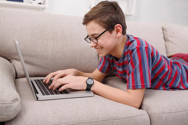 Happy boy with computer on sofa at home. Child engrossed in internet, typing on keyboard