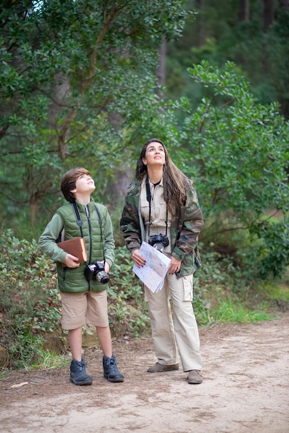 Happy boy and wo with cameras walking in forest. Dark-haired mother and son in coats getting ready to take pictures, looking up. Parenting, family, leisure concept