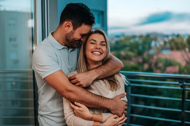 Happy boyfriend and girlfriend standing near window while hugging. 