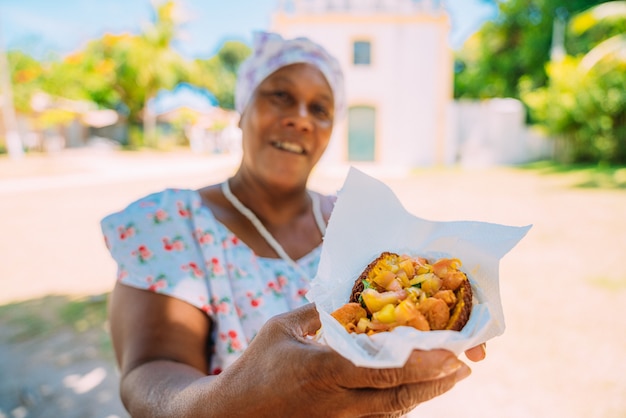 Happy Brazilian woman dressed in the traditional Bahian costume of the Umbanda religion, offering acarajÃ© - typical Bahia food - in the historic center of Porto Seguro in the background