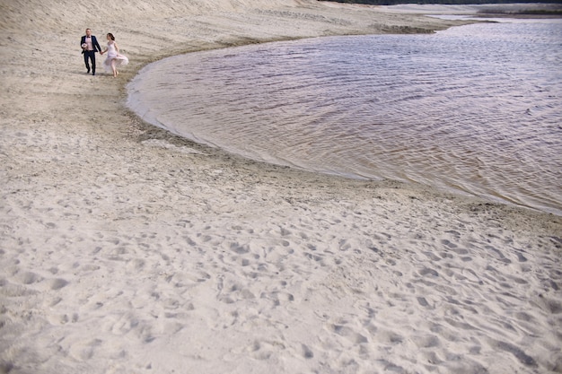 Happy bride and groom run along the shore lake