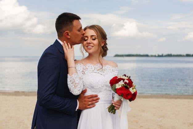 Happy bride and groom walking along the river. romantic walk of newlyweds on sandy beach in sunny day . wedding couple on sea coast . wedding day outdoors. smiling bride and groon huging and kissing