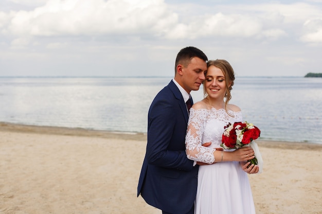 Happy bride and groom walking along the river. romantic walk of newlyweds on sandy beach in sunny day . wedding couple on sea coast . wedding day outdoors. smiling bride and groon huging and kissing