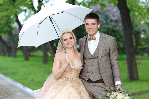 Happy bride and groom with a white umbrella in the rain, in the summer in the park. Open-air wedding.