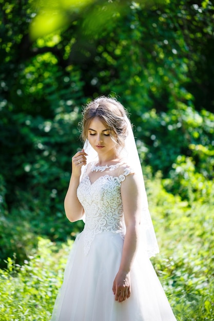 自然の緑の公園で長い白いウェディングドレスとベールで幸せな花嫁。若い女の子の結婚式の画像、女性のメイクや髪型。結婚の概念