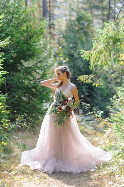 Happy bride in a pink wedding dress. The girl holds a wedding bouquet in her hands. Boho style wedding ceremony in the forest.