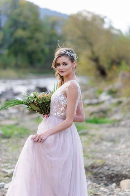 Happy bride in a pink wedding dress. The girl holds a wedding bouquet in her hands. Boho style wedding ceremony near the river.