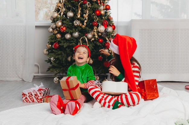 Felice fratello e sorella vestiti da babbo natale sono seduti vicino all'albero di natale a casa nel soggiorno con dei regali. natale