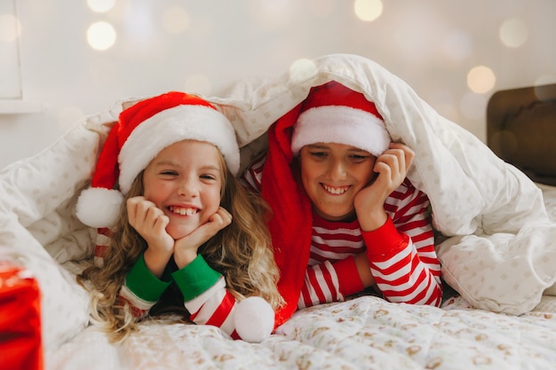 Happy brother and sister in New Year's hats, lie under a blanket in New Year's pajamas. Christmas and New Year.