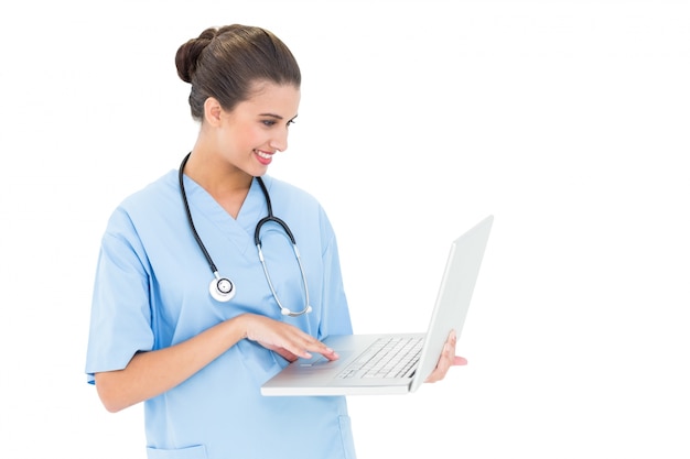 happy brown haired nurse in blue scrubs using a laptop