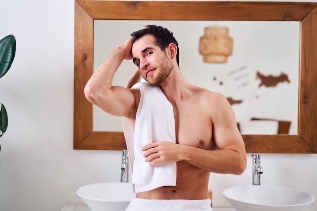 Happy brunet male with towel on shoulder stands near mirror in bath in morning