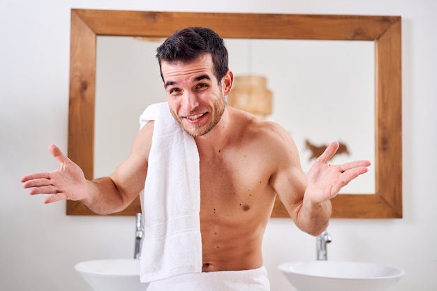 Happy brunet man with towel on his shoulder stands near mirror in morning bath