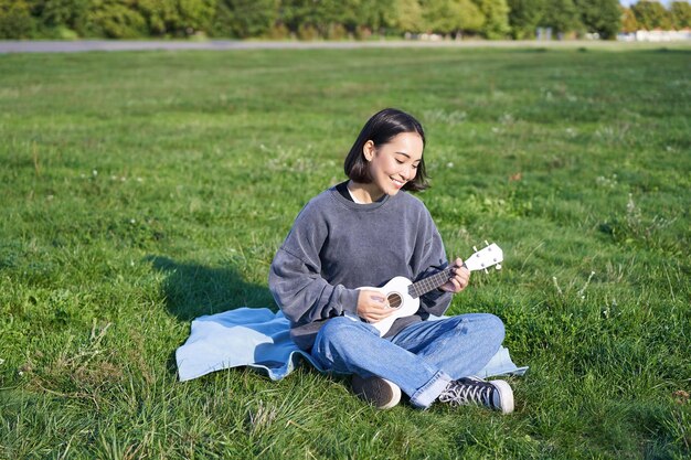 사진 행복 한 갈색 머리 아시아 여자 학생 노래 인생을 편안 하 게 공원에서 잔디에 앉아 우쿨렐레 연주