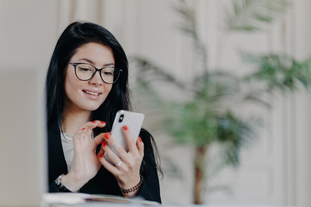 Happy brunette businesswoman uses smartphone wears spectacles sends text messages to partner works on project poses against cozy interior at workplace Technology occupation and work concept