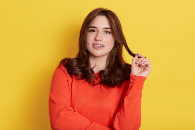 Happy brunette girl flirting and touching her beautiful hair, looking with smile, has romantic mood, wearing casual sweater, isolated over yellow wall.