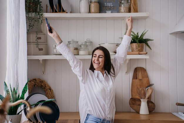 Photo happy brunette girl at kitchen dressed in white shirt and blue jeans raised hands up smiling