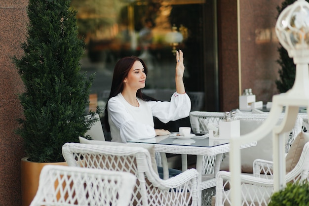 Happy brunette girl sits in a cafe and waves her hand to a friend on the street.