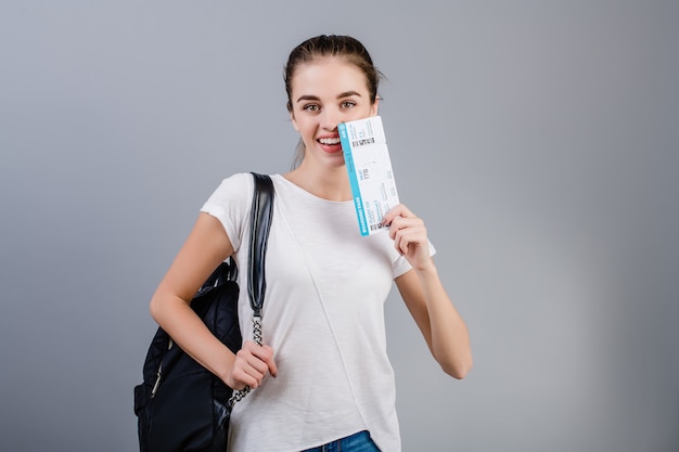 Happy brunette girl with airplane tickets and backpack isolated over grey