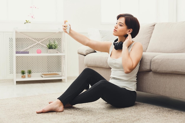 Happy brunette girl with headphones making selfie on smartphone. Young woman having fun sitting on the floor at home