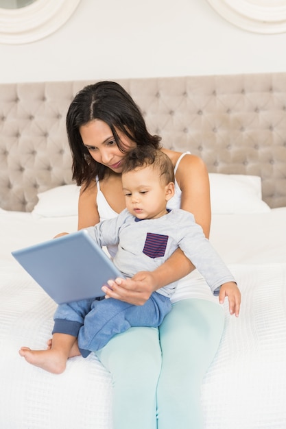 Happy brunette holding her baby and using tablet in bedroom