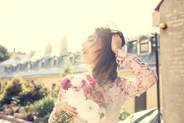 Happy brunette model in pink dress with flowers in hands on a background of city. Vintage toning effect