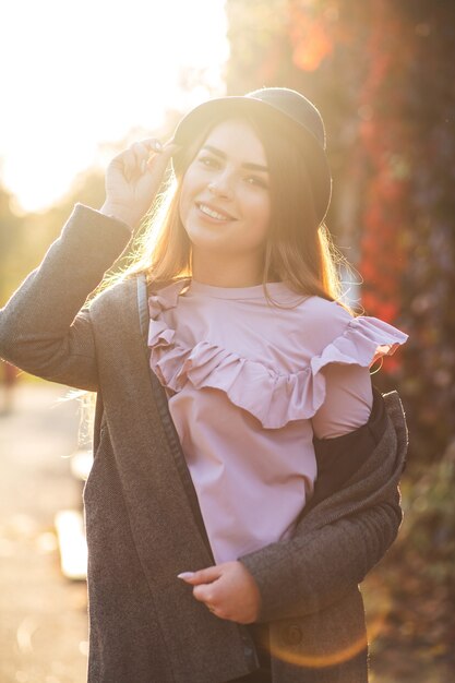 Happy brunette model wearing hat, blouse and coat posing at the sunny street